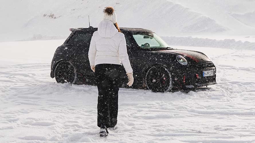 Eine Person geht über eine verschneite Fläche auf einen schwarzen MINI Cooper zu, der vor einer winterlichen Berglandschaft steht.